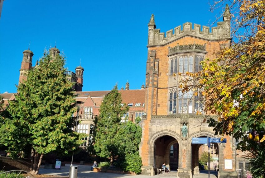 The picture shows the arches of the Newcastle University in the background. In front of the building are trees and a paved road.