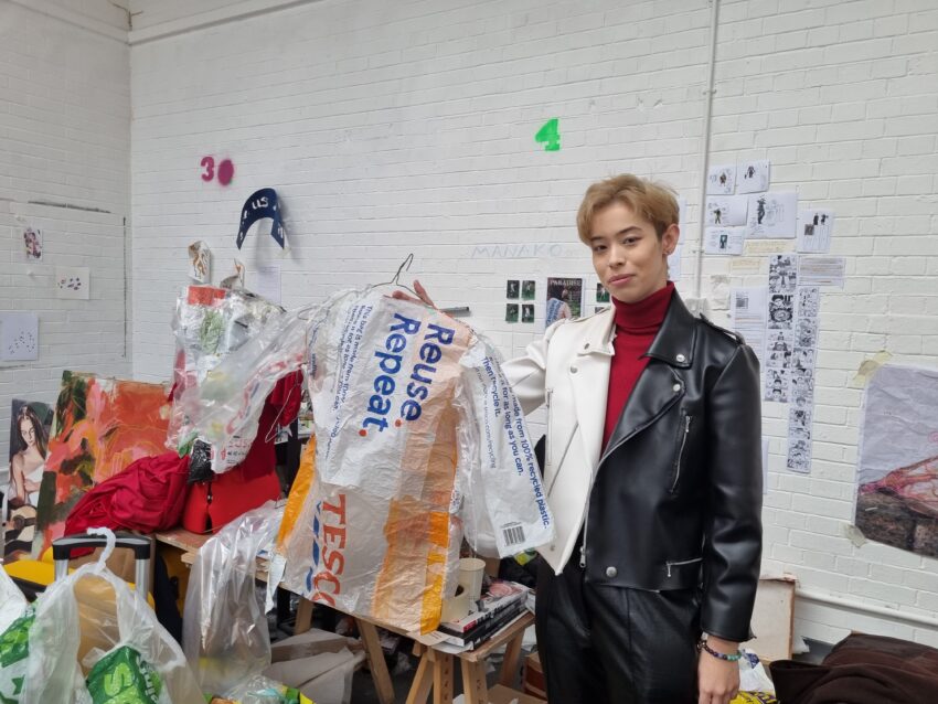 Picture shows Manako Maddison holding a jacket made from plastic bags. In the background is their desk and a white brick wall covered with sketches and posters.