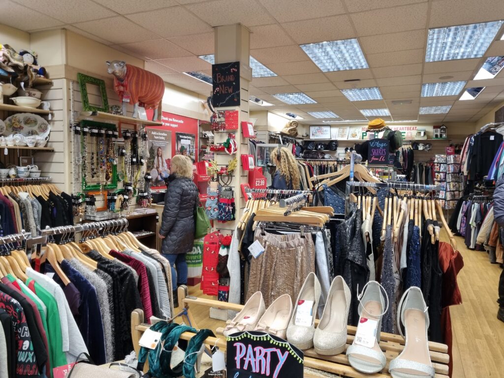 The picture shows the premises of a charity shop. At the forefront of the picture are women's heels, to the left are sweaters on hangers and in the back are more clothes like skirts and shirts on hangers.