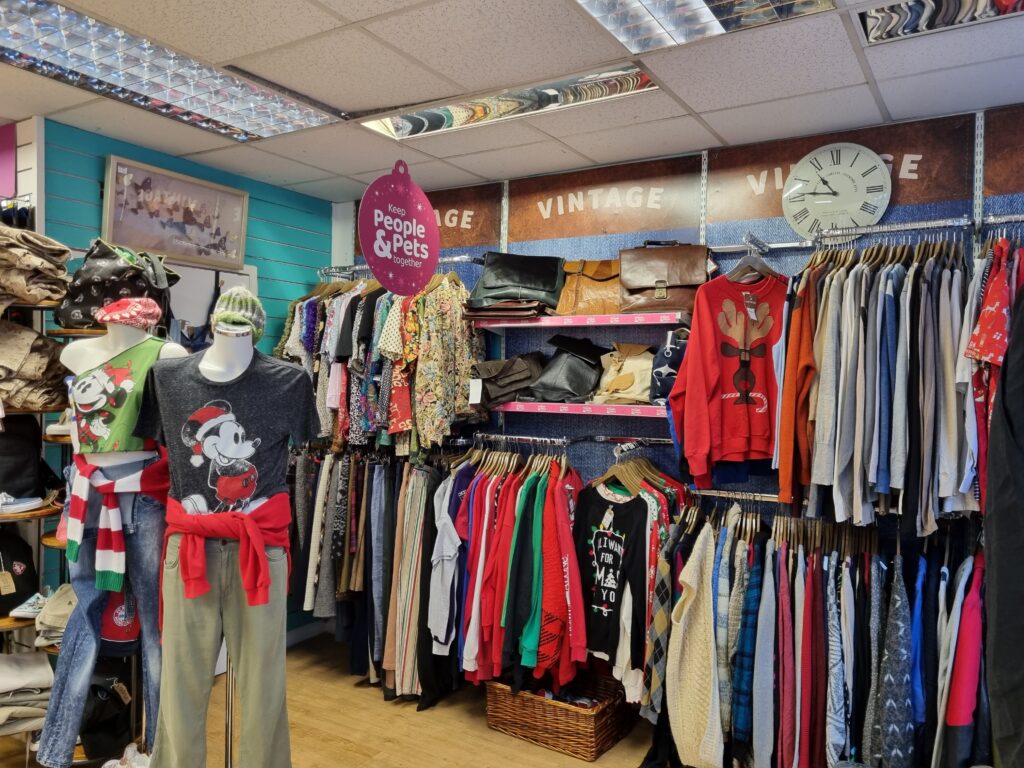 The picture shows the insides of PDSA charity shop. To the left there is a mannequin, wearing a mickey mouse tshirt and green pants. To the back are christmas sweaters on hangers, next to it some other sweaters and shirts.