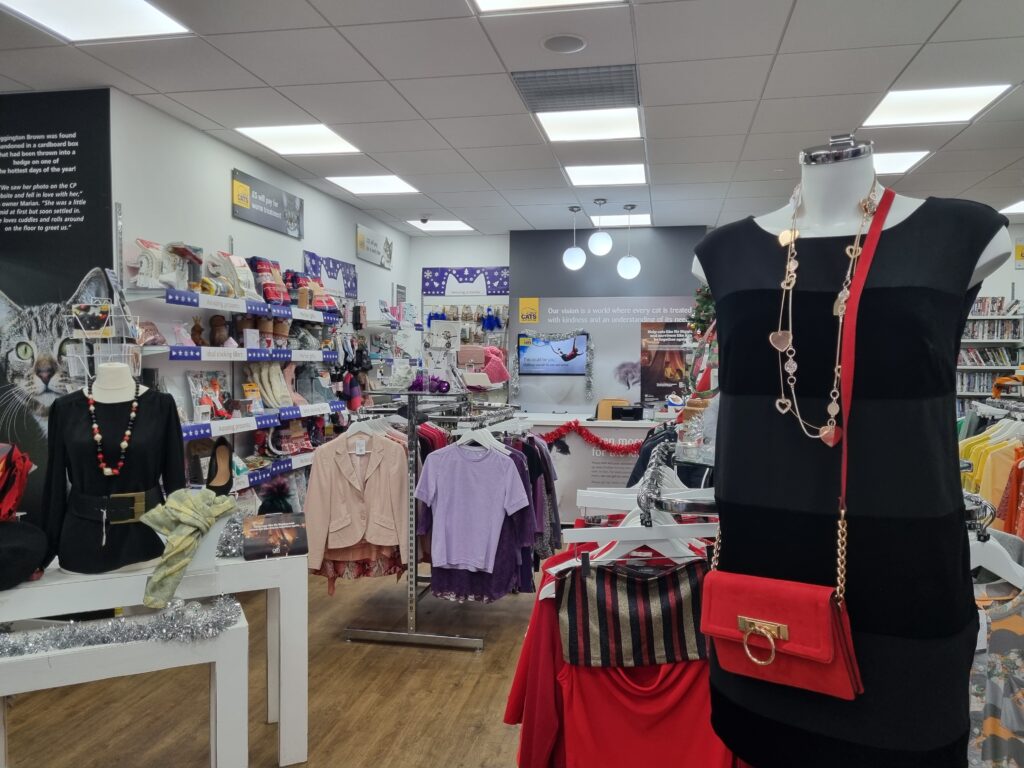 The picture shows the insides of Cats Protection Charity Shop. At the forefront on the right is a mannequin wearing a black dress and small over-the-shoulder red bag. To the end are stalls with shirts and other clothes.