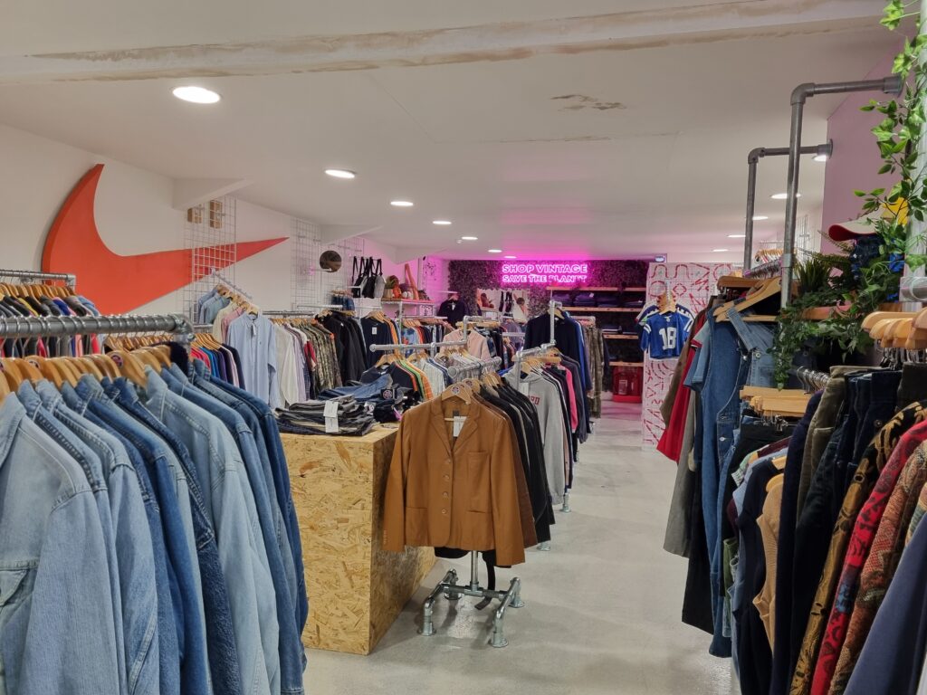 The picture shows the inside of Vintage Scene shop. To the forefront on the left is a stack of denim jackets on hangers. To the back are leather jackets, behind which are some sweaters.