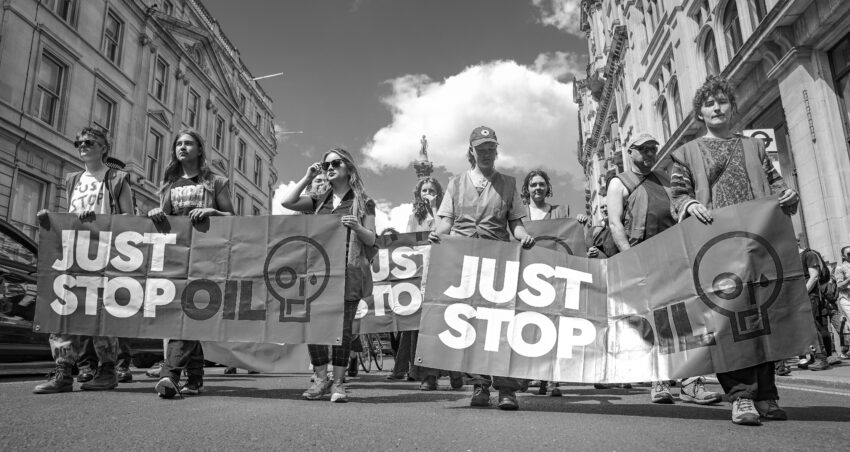 The picture is black and white and shows protesters marching down a street in London. The protesters are holding 'Just Stop Oil' banners.